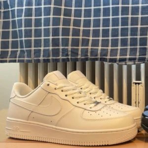 Nike Air Force 1 vita sneakers - Klassiska Nike Air Force 1 sneakers i helvitt skinn med rund tå och platt sula. Skorna har perforerade detaljer på tån, snörning och den ikoniska swoosh-loggan på sidorna. Perfekta för en clean och stilren look.