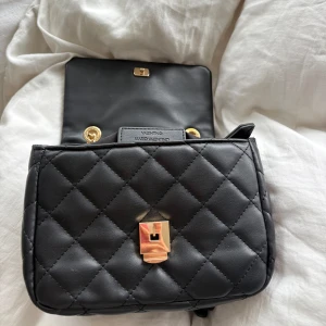 Svart  axelväska från Valentino - Snygg svart axelväska från Valentino med quiltad design, guldfärgade kedjor och detaljer i skinn. Perfekt storlek för det viktigaste. Väskan är i nyskick och endast använd en gång. Fick i födelsedagspresent men tyvärr inte min stil. 