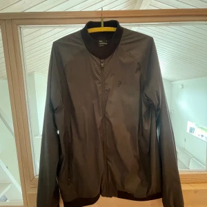 Svart bomberjacka Peak Performance  - Snygg svart bomberjacka från Peak Performance i storlek L. Jackan har klassisk bomberkrage, dragkedja framtill och diskret logga på bröstet. Tillverkad i ett slätt syntetmaterial som känns lätt och smidig. Perfekt för dig som gillar stilren och sportig look.