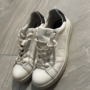 Vita sneakers från Björn Borg - Stilrena vita sneakers från Björn Borg med svart detalj på hälen. Klassisk design med snörning och rund tå. Tillverkade i skinn för en clean look som passar till det mesta. Perfekta för dig som gillar minimalistisk stil.