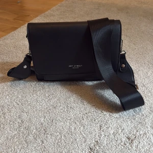 Svart axelväska från Art Street - Snygg svart axelväska från Art Street London med bred axelrem och stilren design. Väskan har ett lock med magnetknäppning och ett extra fack framtill med präglat mönster och har en ljus insida. 