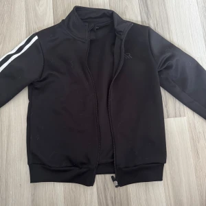 Svart sportig jacka  - Svart sportig jacka i 100% polyester. Jackan har hel dragkedja framtill, ståkrage och vita ränder längs ena ärmen. Perfekt för dig som gillar en clean och enkel stil med en touch av sport.