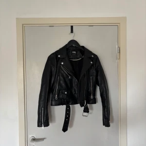 Svart skinnjacka från Zara med bälte - Säljer en svart skinnjacka från Zara med coola dragkedjor, bälte i midjan och klassisk bikerstil. Jackan har flera fickor med dragkedja och detaljer i silver. Perfekt för dig som vill ha en edgy look till vardags.
