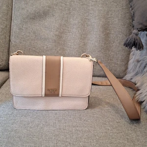 Beige axelväska från Guess - Snygg beige axelväska från Guess med stilren design och justerbar rem. Väskan har en strukturerad yta i syntetmaterial och en bred, ljusbrun rand med Guess-logga framtill. Perfekt storlek för mobil, plånbok och andra essentials.