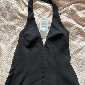 Svart halterneck topp - Snygg svart klänning från zara i halterneck-modell med djup v-ringning. Klänningen har en figurnära passform och är tillverkad i ett slätt material som känns bekvämt mot huden. Perfekt för dig som vill sticka ut med en stilren look. Aldrig använd då den vart för liten. 