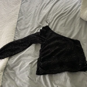 Svart glittrig oneshoulder-top från ONLY - Säljer en svart glittrig oneshoulder-top från ONLY i storlek S. Toppen har lång ärm på ena sidan och är axelbandslös på andra, med snyggt glittrigt mönster i tyget. Aldrig använd. Perfekt festtopp