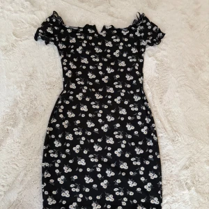 Blommig svart kortklänning från SHEIN - Supersöt svart kortklänning från SHEIN med vita blommor, offshoulder och volangdetaljer vid ärmar och hals. Klänningen har en figurnära passform 