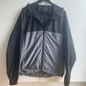 Prada windbreaker - Fysiskt kvitto finns, äkthet garranteras. Nypris 13 000 kr. Säljer en stilren svart och grå vindjacka från Prada med huva och dragkedja. Jackan har en klassisk logga på bröstet och är tillverkad i lätt polyester. Perfekt för dig som vill ha en snygg och funktionell jacka med exklusiv känsla.