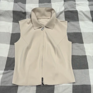 Beige väst med dragkedja och krage - Stilren beige väst med hög krage och hel dragkedja framtill. Västen har en figurnära passform och är tillverkad i ett slätt, mjukt material. Perfekt att styla över en skjorta eller t-shirt för en clean look.