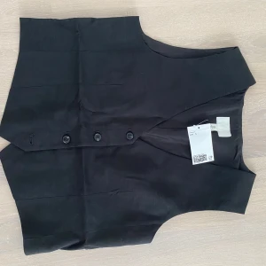 Svart kostymväst med bälte från hm - Stilren svart kostymväst från hm med v-ringning, knäppning framtill med fyra knappar och justerbart bälte i ryggen. Västen har en klassisk, rak passform och är tillverkad i ett slätt tyg som ger en dressad look.