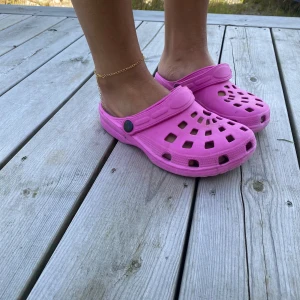 Rosa sandaler i syntetmaterial - Säljer ett par coola rosa sandaler med ventilerande hål och justerbar rem. Skorna är tillverkade i lätt syntetmaterial och har en platt sula som är perfekt för sommardagar. Perfekt för dig som vill ha något bekvämt och färgglatt på fötterna.
