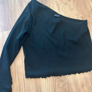 Svart oneshoulder topp Gina Tricot XL - Snygg svart oneshoulder topp från Gina Tricot i storlek XL. Toppen har lång ärm på ena sidan och är croppad med vågig kant nertill. Men skulle säga att det även passar de som har M/L då jag själv brukar ha M .