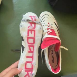 Adidas Predator fotbollsskor vit/röd - Säljer ett par Adidas Predator fotbollsskor i vitt med svarta och röda detaljer. Skorna har snörning, låg profil och tydliga Predator-loggor. Ovandelen är i syntetmaterial och sulan har fasta dobbar för bra grepp på planen. Medföljer originalpåse.
