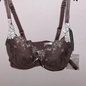 Brun push up-bh med spets från LingaDore - Helt ny.      Superfin brun push up-bh från LingaDore med broderade blommor i vitt och snygga spetsdetaljer längs kanten. Bh:n har justerbara axelband och extra band medföljer. Perfekt för dig som vill ha något både bekvämt och snyggt under kläderna.