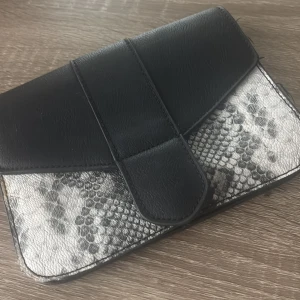 Svart kuvertväska med ormmönster - Stilren svart kuvertväska med lock och detaljer i ormmönstrad syntet. Väskan har flera fack inuti, inklusive ett med dragkedja. Perfekt för dig som vill ha en edgy accessoar med plats för det viktigaste.