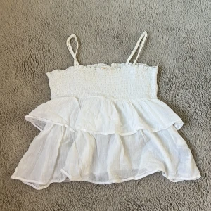 Vit volangig linnetopp med smock - Supersöt vit linnetopp med smala axelband och smockad överdel. Toppen har dubbla volanger som ger en luftig och romantisk känsla. Perfekt för varma dagar och enkel att matcha med jeans eller kjol.