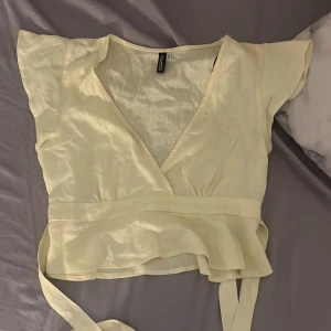 Ljusgul blus - Supersöt ljusgul omlottopp från H&M Divided med v-ringning och knytband i midjan. Toppens material är tunt och flowy, med korta ärmar och volangkant nedtill. Perfekt för dig som gillar en feminin och luftig stil.