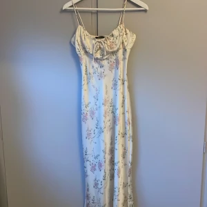 Blommig långklänning från Gina Tricot - Supersöt långklänning med tunna axelband från Gina Tricot. Klänningen är vit med blommigt mönster i rosa och grönt, har en snygg knytning vid bysten och är gjord i ett glansigt, satinliknande material. Perfekt för sommarens alla tillfällen!