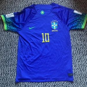 Brasilien Neymar VM 2022 - HELT NY! Snygg blå Brasilien fotbollströja från Nike med Neymar Jr och nummer 10 på ryggen. Tröjan har korta ärmar med leopardmönster i blått och grönt samt gula detaljer. Officiell CBF-logga på bröstet och gröna Nike-loggan. Perfekt för dig som älskar fotboll och Brasilien.