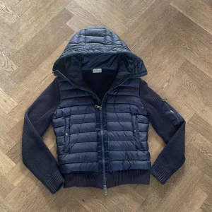 Mörkblå pufferjacka från Moncler - Snygg mörkblå pufferjacka från Moncler med stickade ärmar och huva. Jackan har dragkedja framtill, två fickor med knappar och Moncler-logga på ärmen. Perfekt för kyliga dagar och ger en sportig men clean look. NCF och allt funkar. Perfekt för sommar kvällar🌄🌄