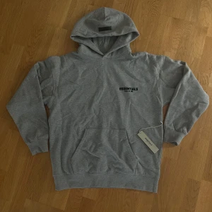 Grå Essentials Fear of God hoodie - Snygg grå hoodie från Essentials Fear of God med svart logga på bröstet och ryggen. Klassisk känguruficka, huva och ribbade muddar. Perfekt för en chill och stilren look. Materialet är mjuk bomullsmix som känns skönt mot huden. Storlek S men passar mer som M