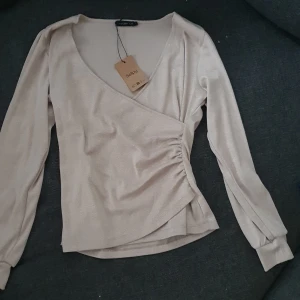 Glittrig omlottopp från STOCKH LM - Snygg beige omlottopp från STOCKH LM med glittrigt tyg och lång ärm. Toppen har v-ringning, lätt rynk på sidan och en figurnära passform. Perfekt för dig som vill sticka ut lite extra med en stilren men ändå festlig look.