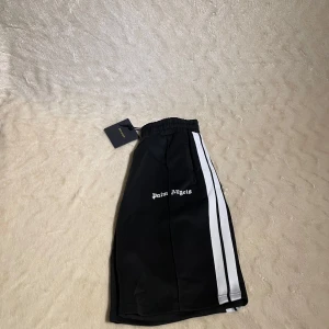 Palm Angels svarta shorts med ränder - Svarta shorts från Palm Angels med vita ränder längs sidorna och logga broderad på ena benet. De har elastisk midja och sportig vibe, perfekta för en avslappnad streetstyle. Materialet känns mjukt och följsamt, troligen polyester.