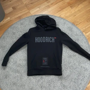 Svart HOODRICH hoodie med tryck - Svart hoodie från HOODRICH med stor logga i grått framtill och röda detaljer. Har huva, känguruficka och tryck på både ärm och huva. Perfekt streetwear-vibe och riktigt skön passform. Materialet känns mjukt och är troligen bomull. Använd 3 gånger typ.