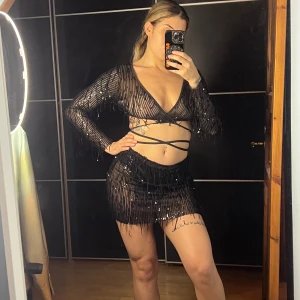 Svart glittrig kortkjol och topp - Säljer en svart kortkjol och topp med glittriga paljetter och fransdetaljer. Djup v-ringning, långa ärmar och snygg snörning runt midjan. Perfekt för dig som vill sticka ut med en festlig look!