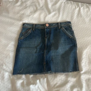 Jeanskjol från Liberto med broderi - Snygg jeanskjol från Liberto i klassisk blå denim. Kjolen är kort med rå kant nertill, coola bakfickor med dekorativa sömmar och ett lila broderat märke med blommor bak. Perfekt för dig som gillar en avslappnad och trendig stil. Midjemåttet är 37cm rakt över