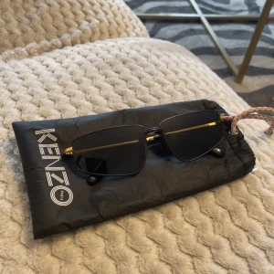 Svarta solglasögon från Kenzo - Snygga solglasögon från Kenzo med rektangulära svarta glas och tunna guldfärgade metallbågar. Stilsäker design med diskret Kenzo-logga på sidan. Kommer med originalfodral. Perfekta för dig som vill sticka ut med en exklusiv vibe.