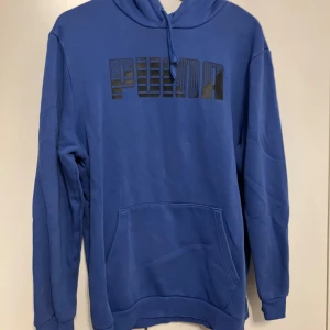Blå hoodie från Puma med logga - Blå hoodie från Puma med stor svart logga framtill. Klassisk känguruficka, huva med snörning och mjukt material. Perfekt för chill dagar eller när du vill ha en sportig look. Långärmad och normal passform.
