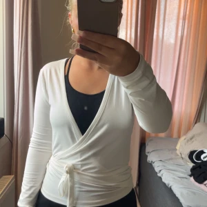 Vit omlottopp med knytband - Snygg vit omlottopp med långa ärmar och knytband i midjan. Toppens v-ringning och figurnära passform ger en clean och stilren look. Perfekt att styla med jeans eller kjol för en fräsch vibe.