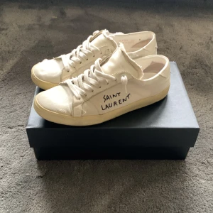 Saint Laurent vita sneakers - Snygga vita sneakers från Saint Laurent med handskriven logga på sidan. Skorna har slitna detaljer och fransade kanter för en cool, avslappnad look. Klassisk rund tå, snörning och platt sula. Perfekta för dig som gillar streetstyle och designer.