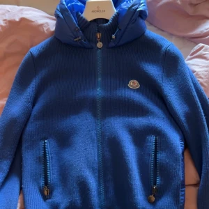 Blå Moncler pufferjacka med huva - Snygg blå pufferjacka från Moncler med stickade ärmar och quiltad kropp. Jackan har dragkedja framtill, två fickor med dragkedja och en vadderad huva. Perfekt för kyliga dagar och har en clean, sportig look med Moncler-logga på bröstet. Ny pris ligger runt 5000kr