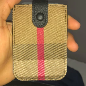 Beige och rutig plånbok med röd rand - Säljer en stilren plånbok i beige med klassiskt rutigt mönster och en röd vertikal rand. Plånboken har svart skinnflik med tryckknapp och plats för flera kort. Perfekt för dig som gillar snygga accessoarer med lite edge.