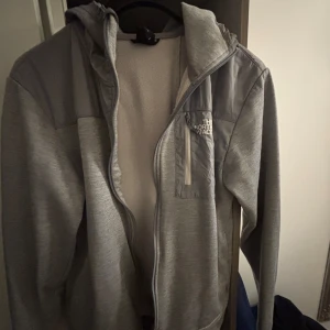 Grå vindjacka från The North Face - Snygg grå vindjacka från The North Face med huva och dragkedja framtill. Jackan har en bröstficka med dragkedja och logga på bröstet. Perfekt för dig som gillar stilren och sportig look. Materialet är mjukt och lätt, med detaljer i syntet.