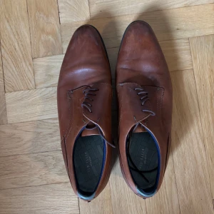 Bruna skinnskor från Ted Baker - Snygga bruna skinnskor från Ted Baker med klassisk snörning och elegant, spetsig tå. Perfekta för dig som vill ha en stilren och tidlös look. Skorna har en tunn sula och diskreta sömmar för en clean finish.
