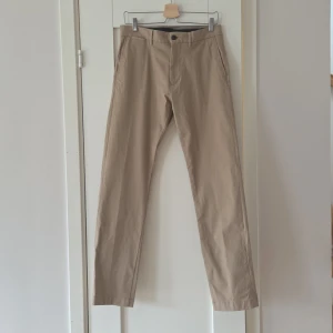 Beiga chinos från Tommy Hilfiger strl.30/34 - Snygga beiga chinos från Tommy Hilfiger med klassisk rak passform. Byxorna har bälteshällor, sidofickor och bakfickor med knapp. Tillverkade i mjuk bomull som känns skön hela dagen. Perfekta för en clean och stilren look.