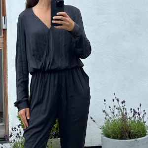 Mörkgrå jumpsuit med lång ärm och v-ringning - Mörkgrå jumpsuit med lång ärm, v-ringad omlott framtill och resår i midjan. Byxorna är raka och har en avslappnad passform. Materialet är mjukt och har ett lätt glansigt fall. Perfekt för dig som gillar en stilren och enkel look.