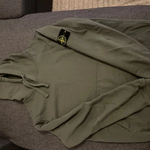 Olivgrön Stone Island hoodie - Säljer en olivgrön hoodie från Stone Island med klassisk patch på vänster ärm. Tröjan har huva med snörning och ribbade muddar. Perfekt för dig som gillar streetwear och vill ha en stilren look.