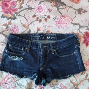 Levi's mörkblå jeansshorts med slitning - Snygga mörkblå jeansshorts från Levi's med råa kanter och slitning på ena benet. Klassisk femficksmodell med dragkedja och knapp framtill. Perfekta för sommaren och ger en cool, avslappnad vibe. 26cm