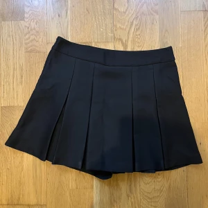 Svart plisserad kortkjol - Snygg svart kortkjol med plisserade veck och bred linning. Det finns en shorts uppsyd under kjol, så man känner sig säker. Materialet känns mjukt och följsamt.