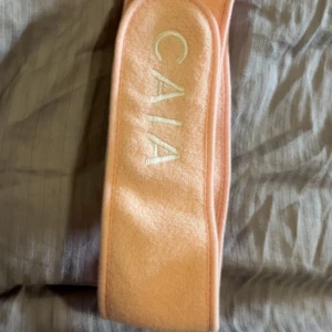 Persikofärgat sminkband från CAIA - Supermjukt och stretchigt sminkband i persikofärgad frotté med CAIA-logga broderad framtill. Perfekt för att hålla håret borta när du sminkar dig eller gör din hudvårdsrutin.