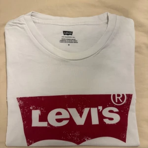 Vit Levi's t-shirt med röd logga - Vit t-shirt från Levi's i bomull med klassisk röd logga framtill. T-shirten har rund halsringning och korta ärmar. Perfekt för en clean och avslappnad stil.