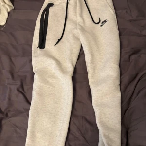 Nike tech byxor - Snygga vita mjukisbyxor från Nike med svart logga på låret, svarta dragsnören i midjan och en cool dragkedjeficka på sidan. Sköna och stilrena, perfekta för chill eller träning. Tillverkade i mjukt bomullsmaterial. Välldigt sköna men har blivit för snå för mig. Köpta från nike.com.