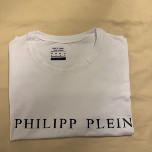 Vit Philipp Plein t-shirt i bomull - Snygg vit t-shirt från Philipp Plein med klassisk rund hals och svart logotyptryck framtill. Tillverkad i mjuk bomull som känns skön mot huden. Enkel och stilren design som passar perfekt till jeans eller shorts.