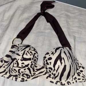 Zebramönstrad bikiniöverdel LingaDore - Snygg bikiniöverdel från LingaDore med svartvitt zebramönster och vadderade kupor. Halterneck med breda mörkbruna knytband och guldfärgad ringdetalj. Tillverkad i mjuk nylonblandning med polyesterfoder. Perfekt för strandhäng och poolparty.