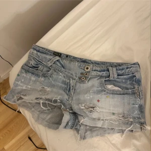 River Island ljusblå jeansshorts - Ljusblå jeansshorts från River Island med slitna detaljer, råa kanter och flera knappar framtill. Shortsen har klassiska fickor, små färgprickar och en cool leopardmönstrad patch bak i midjan. Perfekta för en avslappnad sommarstil.