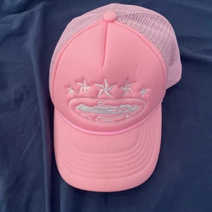 Rosa truckerkeps från Corteiz - Snygg rosa truckerkeps från Corteiz med vit brodyr framtill och på sidorna. Kepsen har mesh-bakstycke för extra ventilation och justerbar snapback. Broderade stjärnor och logga ger en unik vibe. Perfekt accessoar för att lyfta din streetwear-look.
